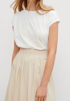 Kvinde iført en hvid kortærmet top med subtile perlemønstre, stoppet ned i et beige plisseret nederdel med høj talje.