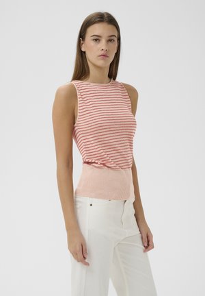 Junge Frau trägt ein ärmelloses pink-rot gestreiftes Top und weiße Hose, steht vor einem schlichten hellen Hintergrund.