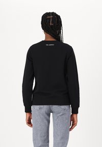 Felpa nera con maniche lunghe, texture morbida, scollatura rotonda e logo bianco "KARL LAGERFELD" sulla schiena. Abbinata a jeans in denim chiaro.