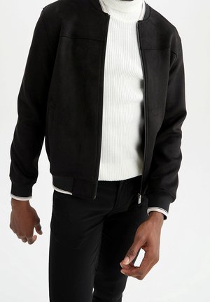 Man die een zwart bomberjack met ritssluiting draagt over een witte geribbelde coltrui en zwarte broek, staand tegen een witte achtergrond.