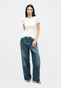 Femeie cu păr drept și întunecat, purtând un top alb cu mâneci scurte, blugi albaștri cu picioare largi și sandale negre cu degete dezvelite, stând pe un fundal alb.