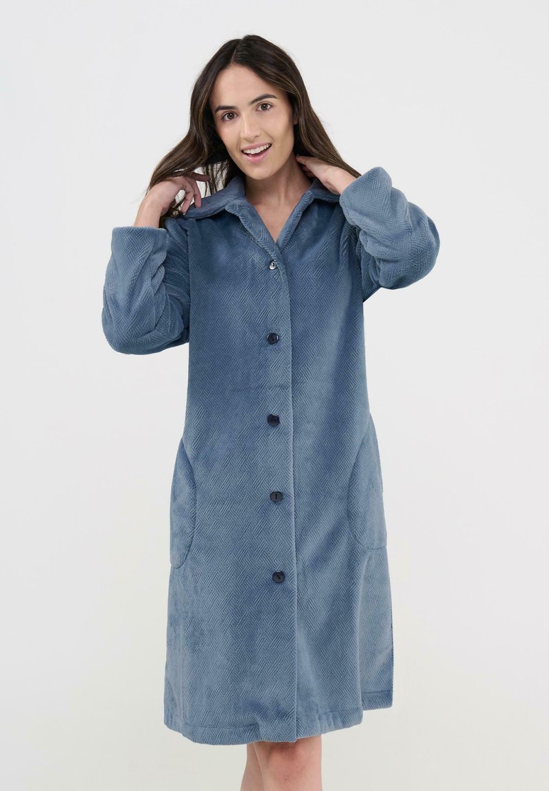 Mujer con cabello largo y marrón, vistiendo un abrigo azul con textura hasta la rodilla y botones, con bolsillos, ajustándose el cuello y sonriendo.