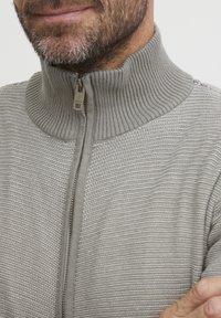 Grauer Zip-Hoodie mit geripptem Kragen. Der strukturierte Stoff weist horizontale Streifen und ein markantes Reißverschlussdetail auf.