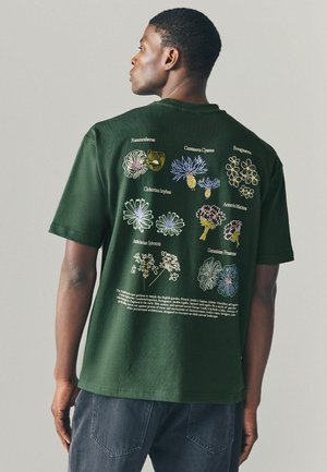 Mørkegrøn bomulds T-shirt med farverig blomsterbroderi på bagsiden, rund hals og korte ærmer. Teksten inkluderer plante-navne og beskrivelser.