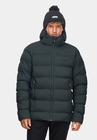 Ciemnozielona kurtka puchowa z wysokim kołnierzem, zamkiem z przodu i dwiema bocznymi kieszeniami. Brzegowa czarna czapka z logo spoczywa na głowie modela.