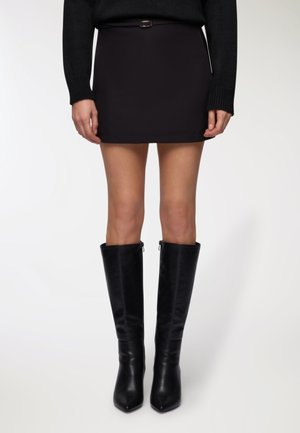 Donna che indossa una gonna mini nera, un maglione di maglia nera e stivali in pelle nera alti fino al ginocchio, in piedi contro uno sfondo bianco.