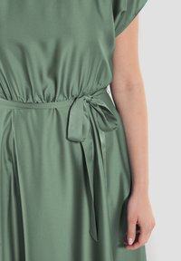 Grön sidentopp med avslappnad passform, rynkad midja och satinrosett på sidan. Slät textur och flödande design med korta ärmar.