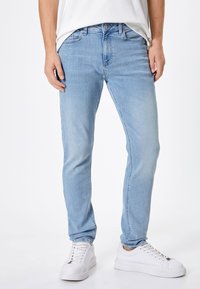 Jeans de mezclilla azul claro con corte slim, que cuentan con cinco bolsillos y un cierre de botón. Combinados con zapatillas blancas y una camiseta blanca lisa.