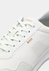 Adidași albi cu un superior din pielefină, detalii perforate pe lateral, șireturi tonale și un branding subtil în aur. Prezintă o talpă din cauciuc texturat.