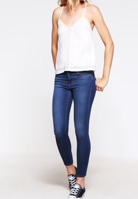 Top sottile bianco con pizzo, abbinato a jeans skinny blu scuro e sneakers nere basse. L'outfit presenta spalline sottili e un design aderente.