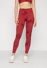 Kobieta ubrana w czerwone legginsy w tie-dye z wysokim stanem oraz pasujący stanik sportowy, w połączeniu z białymi butami sportowymi, stojąca na jednolitym tle.