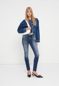 Ülepäine denim-jakk, helesinised teksad ja mustad teravad kontsad. Mudel hoiab valget käekotti rihmaga. Lihtne, puhas disain.