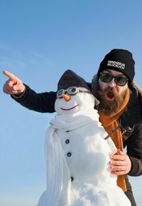 Snowman with a carrot nose, wearing a brown aviator hat, round glasses, and a white scarf, next to a person in a black beanie and sunglasses.