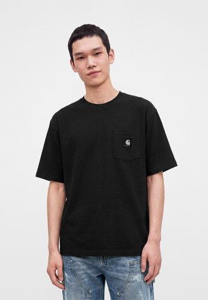 Giovane uomo che indossa una t-shirt nera Carhartt con tasca e jeans azzurri usurati, in piedi davanti a uno sfondo chiaro uniforme.