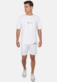 T-shirt bianca in cotone con maniche raglan e stampa nera del logo, abbinata a pantaloni corti bianchi e sneakers bianche con dettagli rossi.