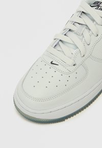 Heled hall nahkist sneaker, millel on perforatsioonidega varvaste osa, lame pael ja must Nike logo. Sisaldab läbipaistvat kummist väliskesta.