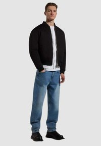 Giubbotto bomber nero con zip e orlo a costine, abbinato a jeans chiari scoloriti e scarpe nere. T-shirt bianca sotto.