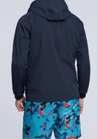 Námořnicky modrá nepromokavá bunda s kapucí, s předním zipem a elastickými manžetami, nošená přes veselou vzorovanou plavkové šortky.