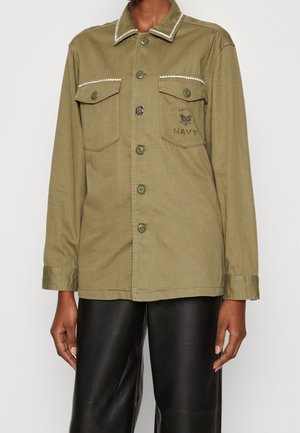 Olive green buttoned jacket with two chest pockets and white trim on collar and pocket flaps, worn with black pants.