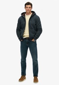 Man in een marineblauwe hoodie met rits en een bruine fleece voering, een crèmekleurige trui, blauwe jeans, bruine leren laarzen en een polwatch, staande met handen in zijn zakken.