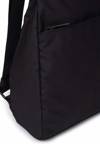 Bolsillo lateral de mochila de tela negra con pestaña del tirador de la cremallera parcialmente visible y correa de hombro ajustable unida al lateral.