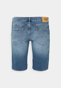 Farkkushortsit vaaleansinisessä pesussa, takaa kuvattuna, joissa on kaksi taskua ja brändätty vaaleanruskea nahkalappu vyötäröllä.