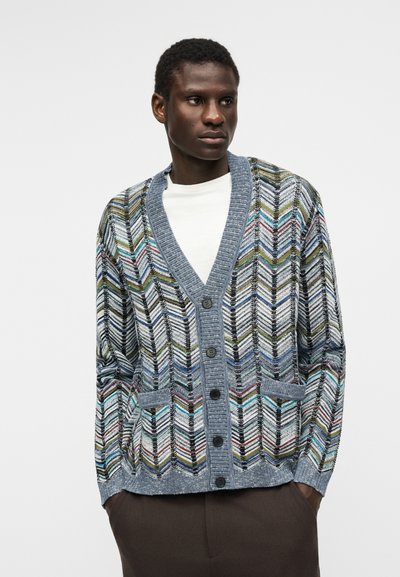 Άνδρας που φορά πολύχρωμη ζακέτα με ζιγκ-ζαγκ μοτίβο και μπλε ρίγα πάνω από λευκό πουκάμισο, στέκεται με τα χέρια στις τσέπες.