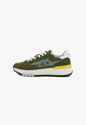 Oliijfgroene sneaker met witte veters, grijs bliksemvormig ontwerp, zwarte geometrische symbolen, witte hiel, gele accenten en zwarte zool.