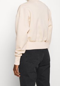 Beżowa bluza z wysokim kołnierzem, luźny krój oraz ściągacze przy rękawach i u dołu, w zestawie z ciemnymi dżinsami. Gładka tekstura materiału.