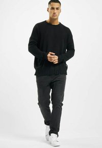 Pull en tricot noir avec texture côtelée et ourlet effiloché, porté avec un jean foncé et des baskets blanches, montrant une silhouette décontractée et ajustée.