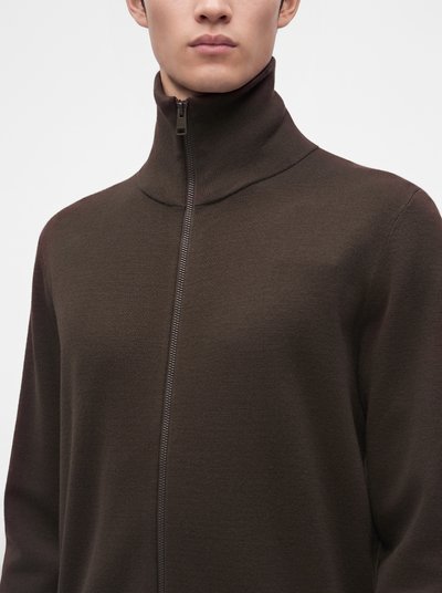 Homme portant un pull marron foncé à col montant zippé, la fermeture éclair complètement fermée, sur un fond uni.