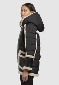 Sort polstret jakke med beige kunstpelskant, der har hætte, frontlynlås og sidelommer. Glat tekstur med en struktureret design.