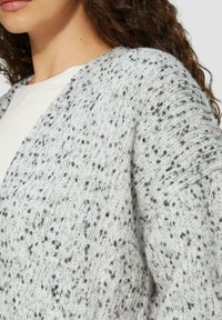 Lichtgrijze cardigan gemaakt van gestructureerde brei-stof met zwarte spikkels, met een open voorkant en losse mouwen.