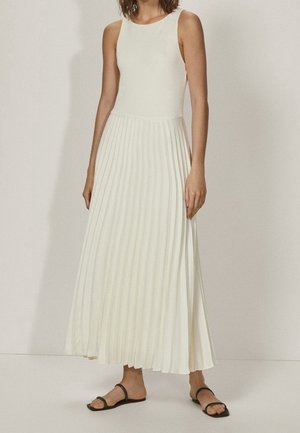 Sleeveless white maxi dress with fitted top and pleated skirt worn with black flat sandals.