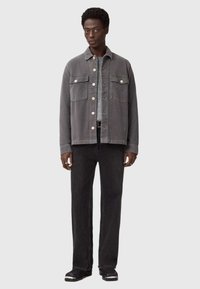 Grijze katoenen jas met twee borstzakken, knoopsluiting, lange mouwen en een relaxte pasvorm, gecombineerd met zwarte jeans en schoenen.