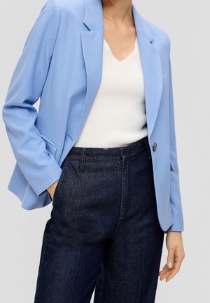 Person iført lyseblå blazer over hvid top med mørkeblå høj talje jeans, én hånd i lommen, stående mod en ensfarvet baggrund.