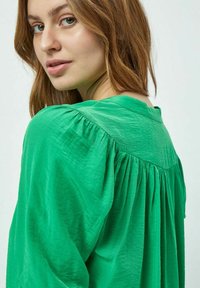 Blusa verde con vestibilità morbida, caratterizzata da dettagli arricciati sul retro, texture morbida e scollatura rotonda. Design minimalista.