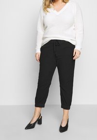 Biały dziergany sweter z dekoltem w serek, w zestawie z czarnymi zwężanymi spodniami z elastycznym pasem i wiązaniem z przodu oraz czarnymi butami na obcasie.