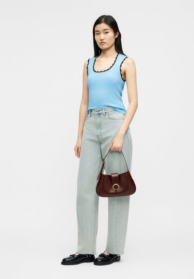 Γυναίκα που φοράει ανοιχτό μπλε αμάνικη μπλούζα με μαύρη επένδυση, τζιν ανοιχτού χρώματος, μαύρα μοκασίνια, κρατώντας σκούρα καφέ τσάντα χειρός με χρυσό δαχτυλίδι ως διακοσμητικό στοιχείο.