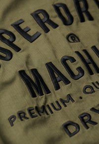 Olivenfarvet stof med et fiskebensmønster, prydet med fed broderet tekst i sort, der lyder "SUPERDRY MACHINERY PREMIUM QUALITY DRY."