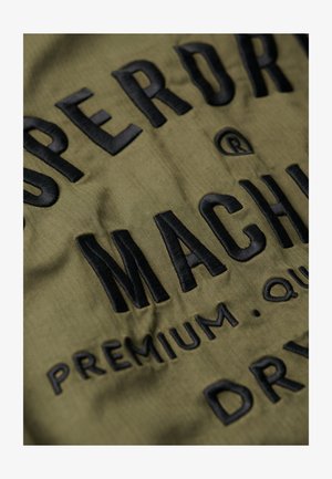 Olivengroen stof met een visgraatstructuur, met dikke zwarte geborduurde tekst die leest: "SUPERDRY MACHINERY PREMIUM QUALITY DRY."