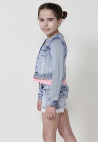 Jeans jakna s svetlo modrim pranjom, okrašena ovratnikom i ružičastim detaljima, u kombinaciji s traper šortsevima s čipkastim rubom.