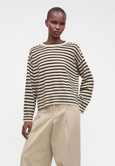 Femme portant un haut à manches longues rayé noir et blanc et un pantalon beige ample, debout devant un fond clair uni.