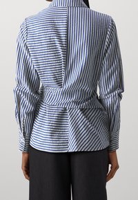 Camicia a strisce blu e bianche con colletto, maniche lunghe e vita con cinturino. Presenta un taglio preciso e un tessuto texturizzato.