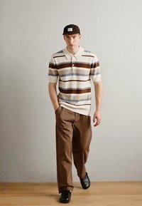 Kortermet polo i kremfarge med brune og blå striper, sammen med løse brune bukser og svarte sko. Teksturert strikk med krage.
