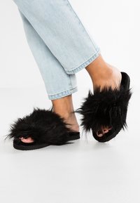 Black fur-slide slippers with a fluffy upper, a flat black sole, and an open-toe design, worn with light blue cuffed jeans.