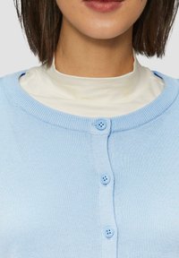 Lichtblauwe cardigan met een ronde halslijn, voorzien van zes knopen aan de voorkant en een gestructureerde brei. Draagt over een witte turtleneck.
