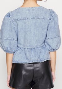 Kvinna som bär en ljusblå denimblus med puffärmar och svarta lädershorts, sedd bakifrån mot en vit bakgrund.