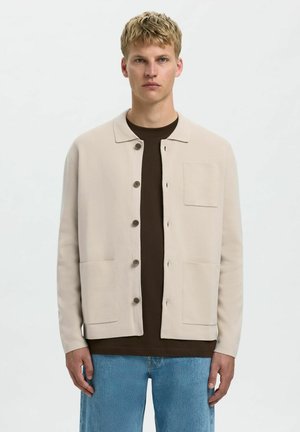 Beige knit cardigan with a collar, front buttons, two pockets, and a textured finish, layered over a dark brown t-shirt and light blue jeans.