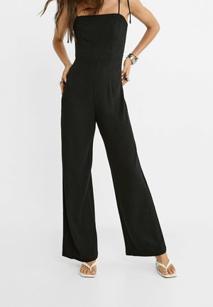 Femme portant une combinaison noire sans manches à jambes larges et des sandales tongs blanches, debout les mains sur les hanches.